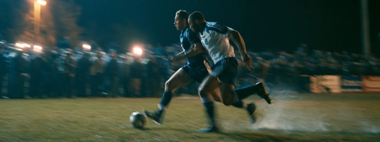 Deux footballeurs courant côte à côte vers le ballon, tribunes éclairées en nocturne